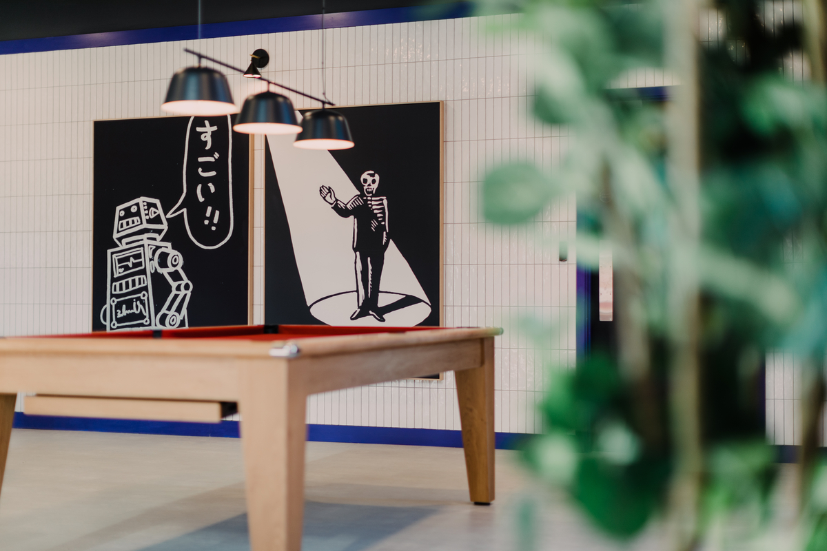 A modern room with a pool table and two large graphic artworks on the wall. The left artwork features a robot with Japanese text, while the right depicts a man in a suit under a spotlight. A leafy plant is partially visible in the foreground.
